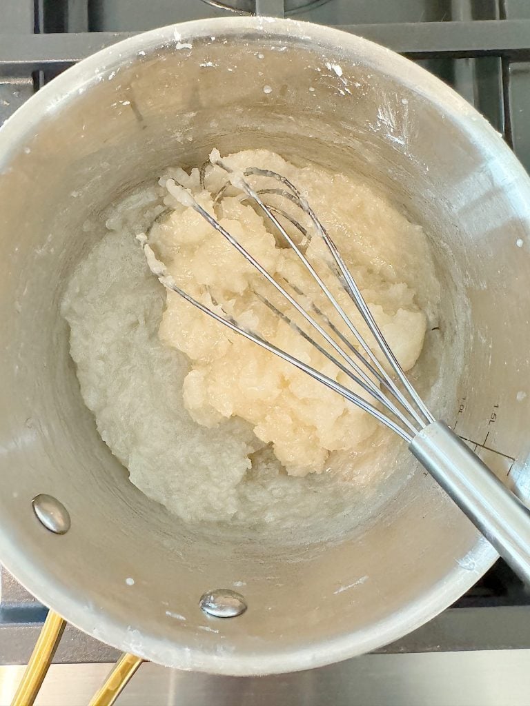 A pot containing a thick, white mixture being stirred with a whisk on a stove.