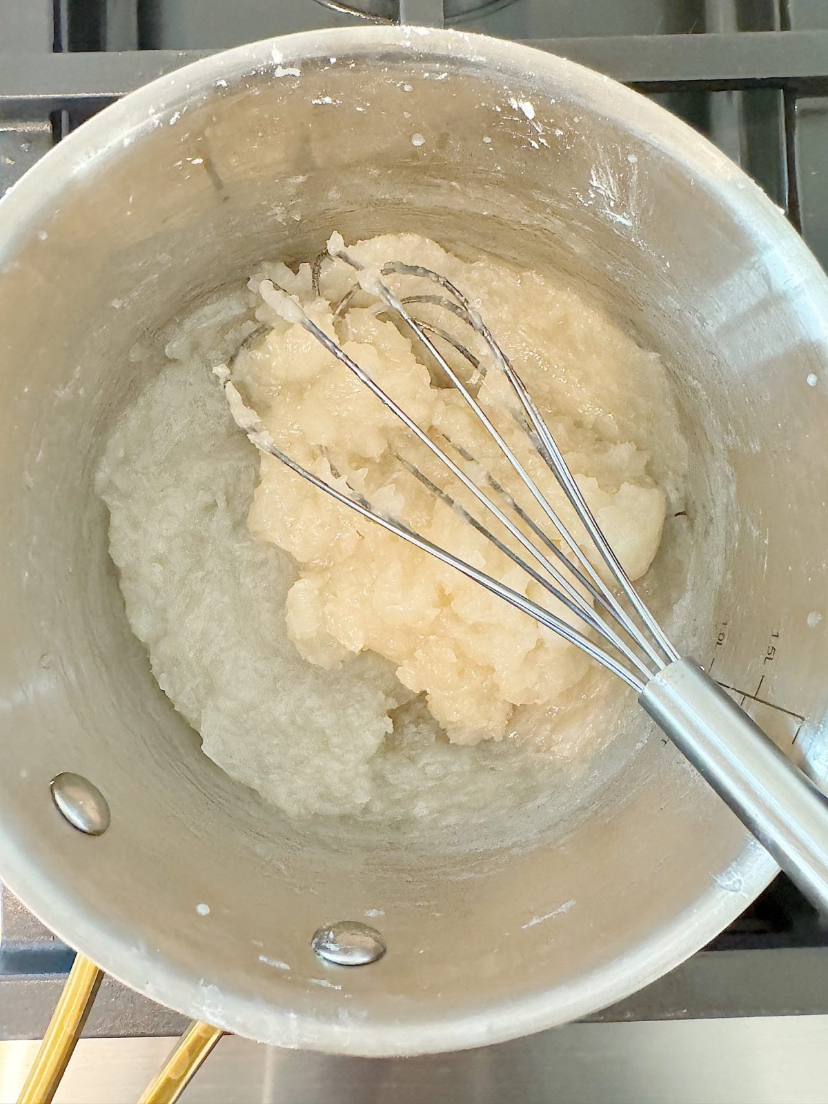 A pot containing a thick, white mixture being stirred with a whisk on a stove.