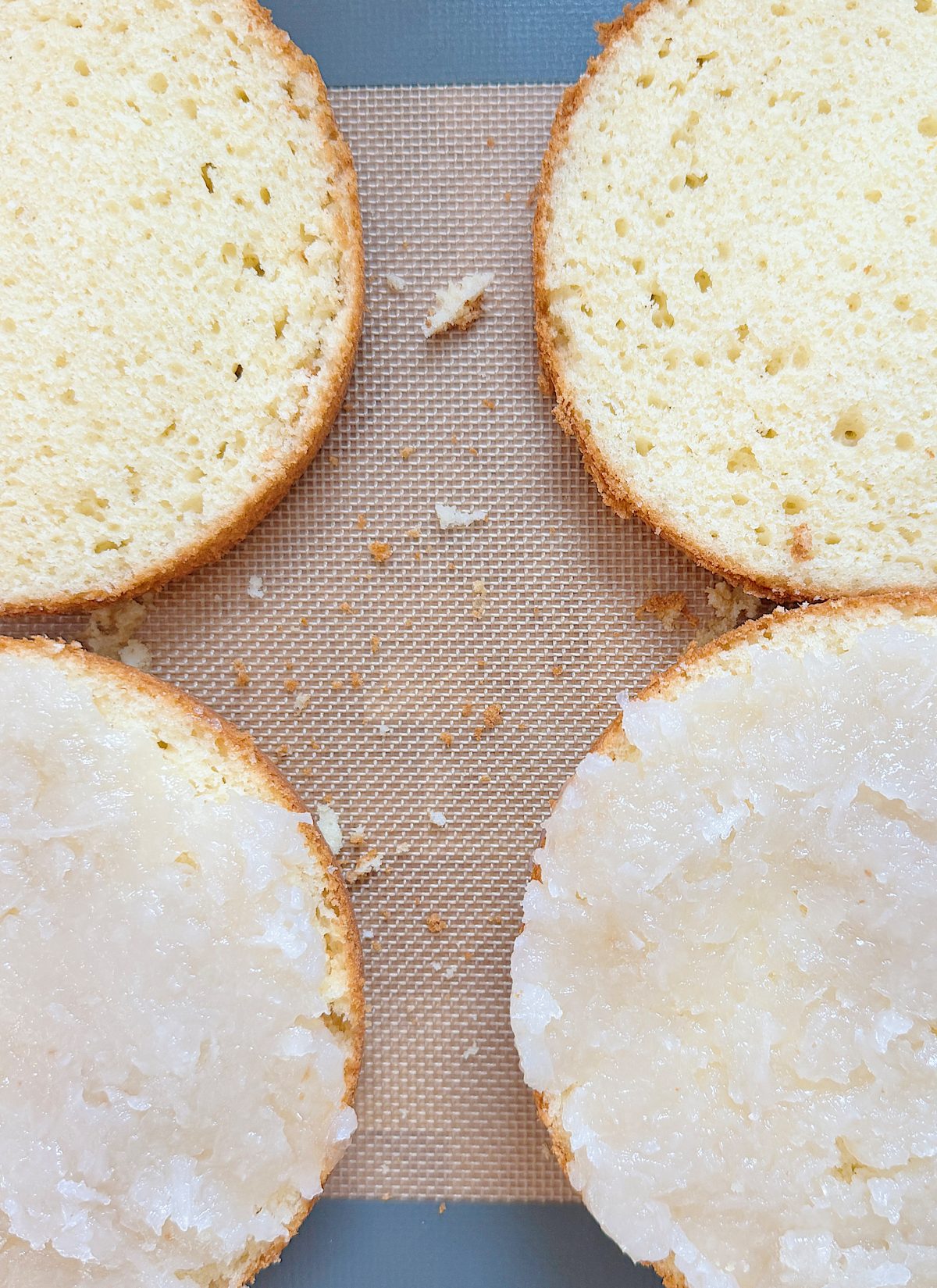 Four cake layers on a baking mat, with two plain and two covered in a white frosting. Crumbs are scattered on the mat.