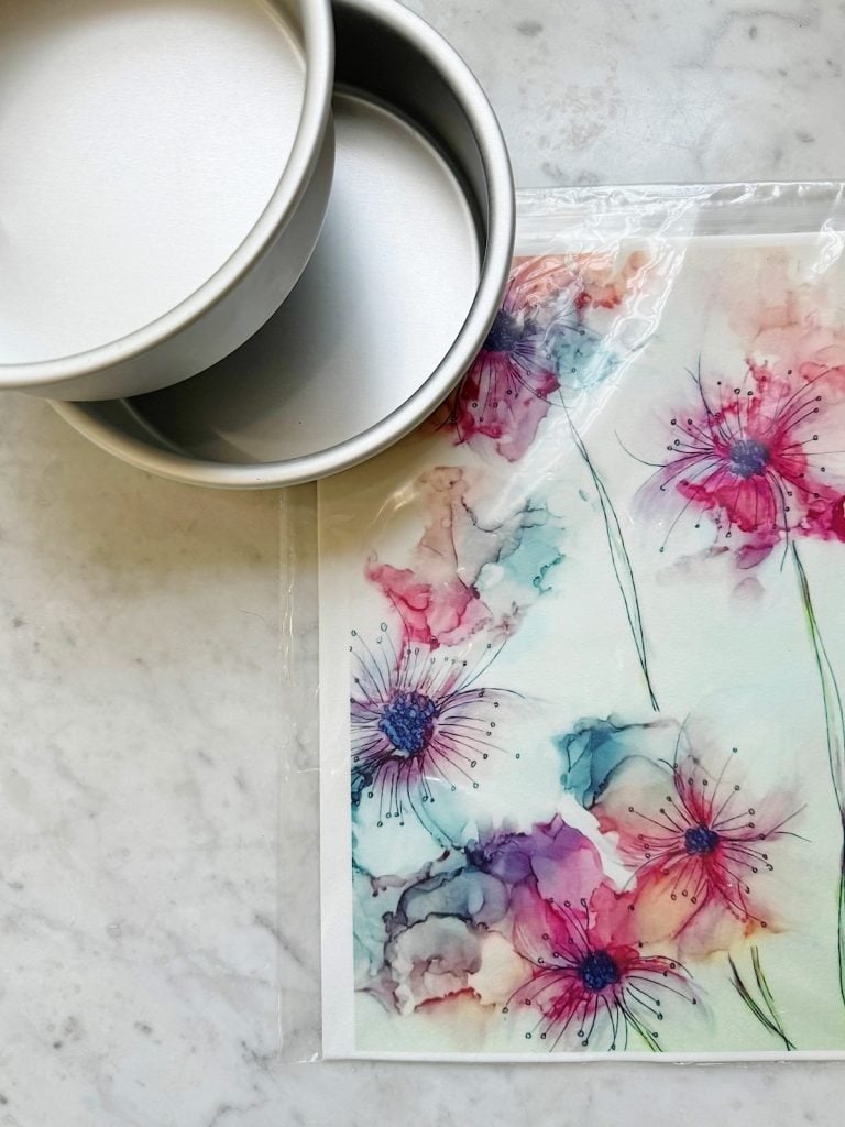 Two nested round baking pans sit on a marble surface alongside a watercolor print featuring vibrant pink, purple, and blue flowers.