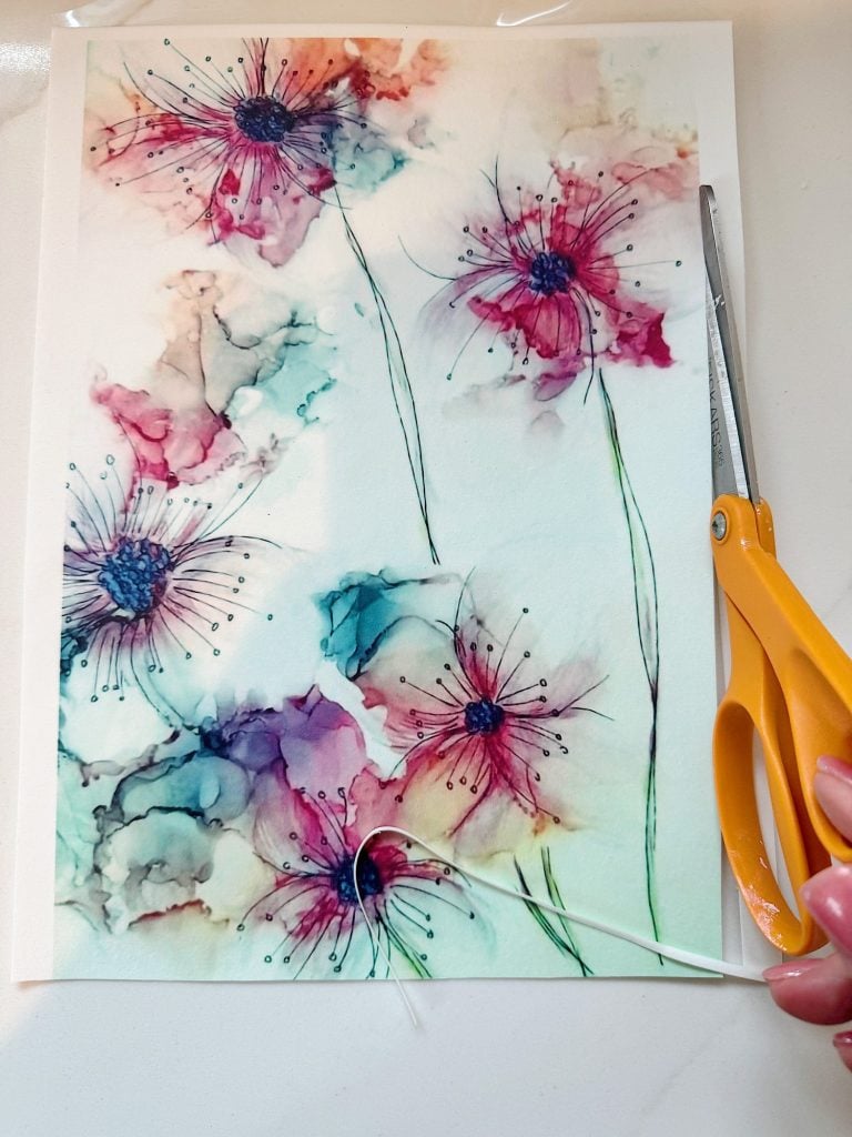 A hand holds a pair of scissors over a watercolor painting of abstract flowers in shades of pink and blue on white paper.