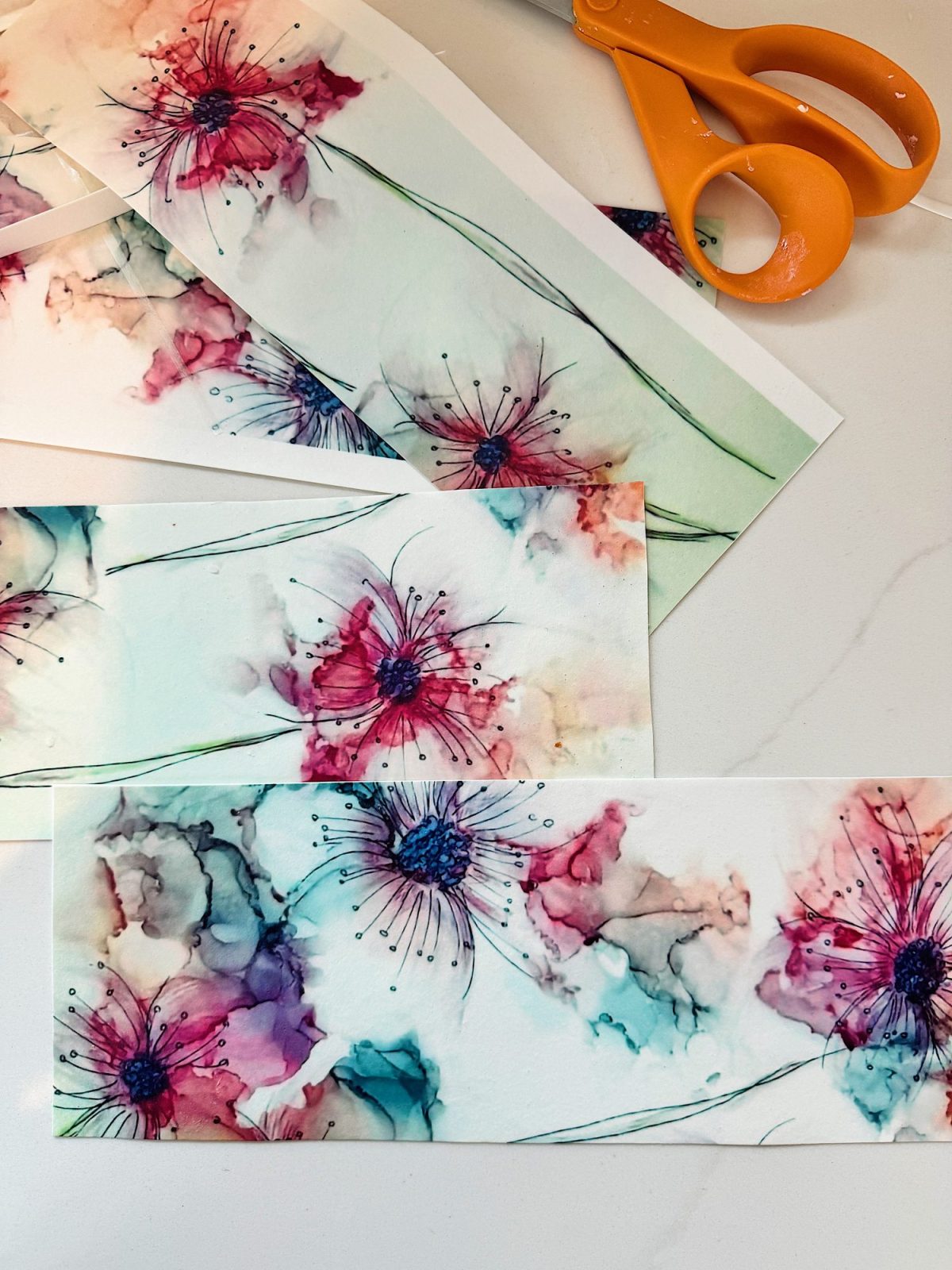 A pair of orange scissors on a white surface beside several watercolor floral paintings featuring vibrant red, pink, and teal flowers.