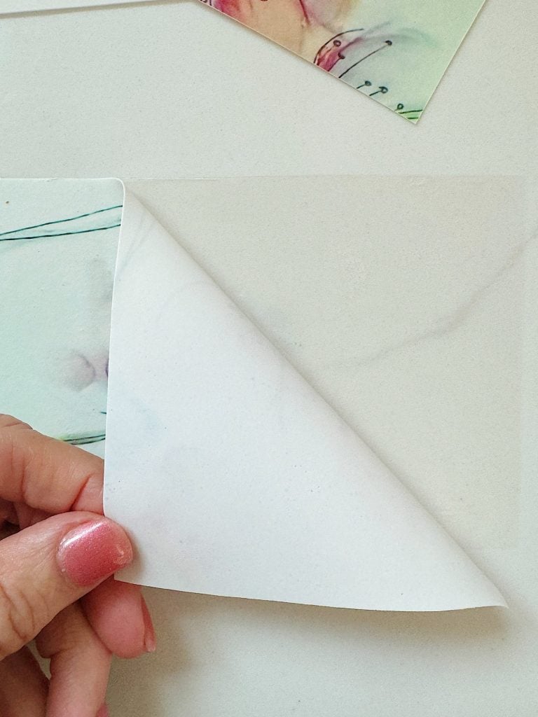 A hand with pink-painted nails peels a corner of a piece of paper away from a white surface, revealing the back side of the paper.