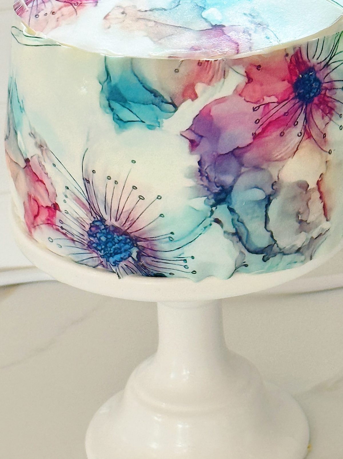 A white cake on a pedestal stand decorated with watercolor-style edible flowers in shades of pink, blue, and purple.