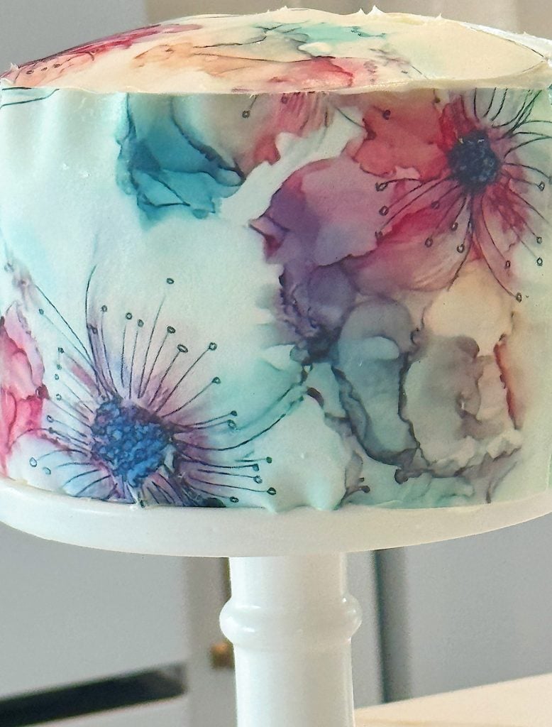 A white cake on a white stand decorated with watercolor-style floral designs in shades of blue, pink, and purple.