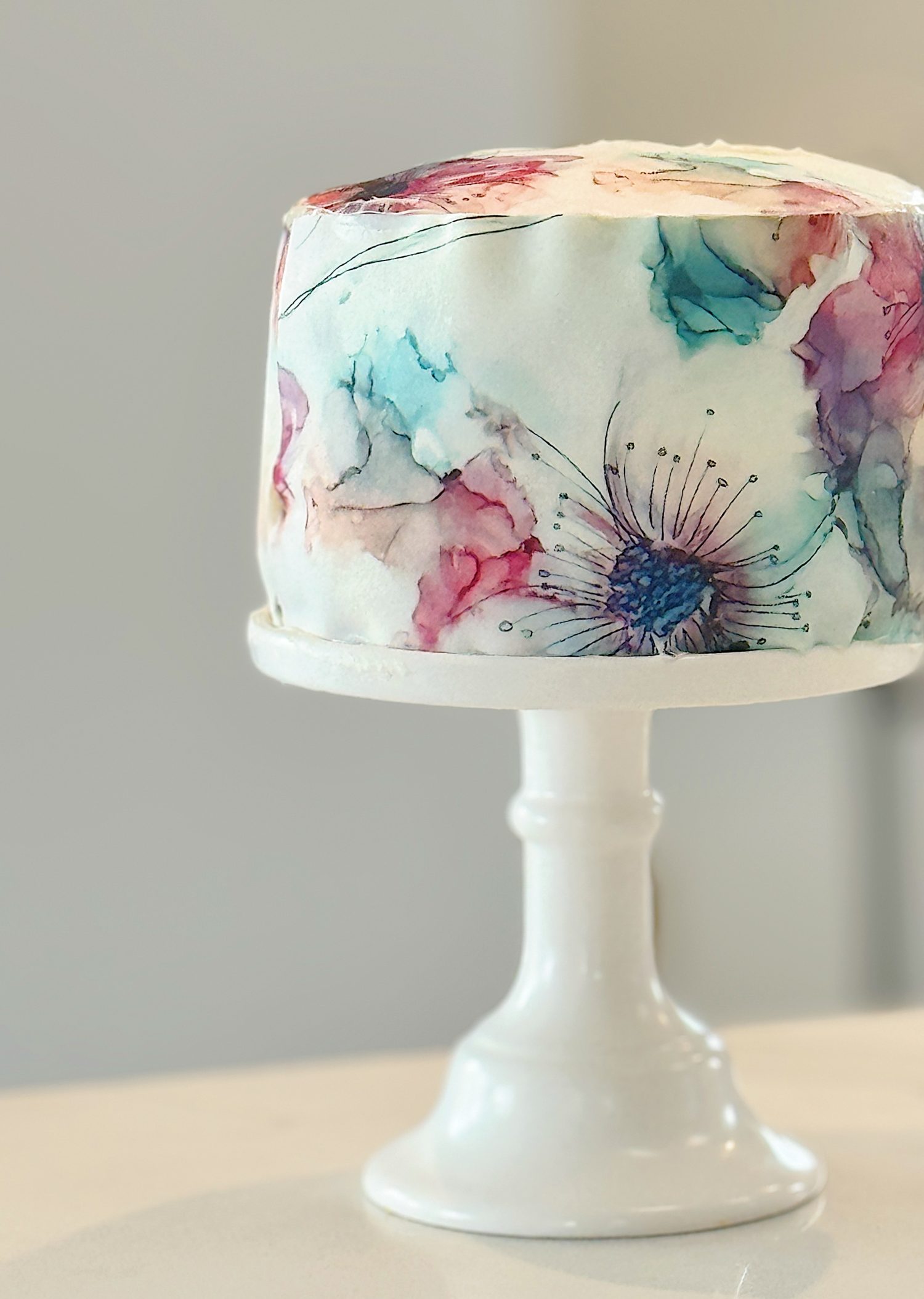 A white frosted cake with watercolor-style floral designs in various colors sits on a white cake stand.