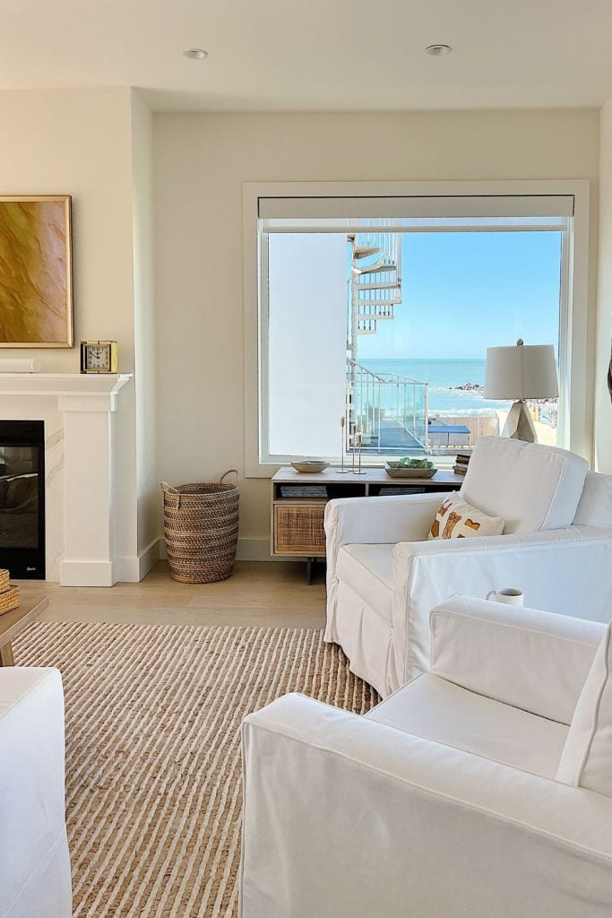 A bright living room with white furniture, a striped rug, a fireplace, and a large window offering a view of a spiral staircase and the ocean outside. Perfect for enjoying peaceful moments or unwrapping Mother's Day gifts from Amazon amidst serene surroundings.