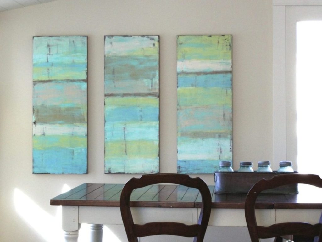 Three elongated abstract paintings with blue and green tones, reminiscent of Willamette Valley wineries, hanging on a white wall above a dining table with chairs.