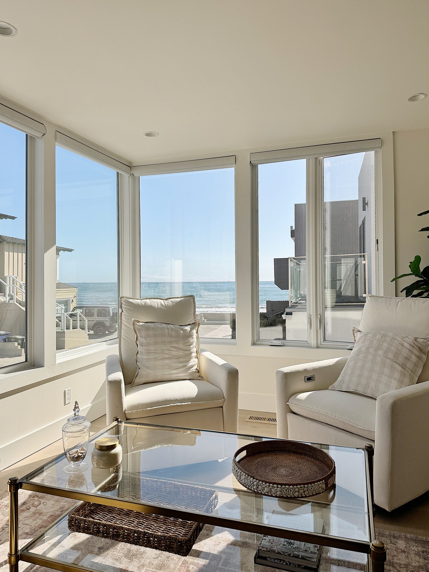 A bright sunlit living room with large windows overlooking a beach. The room includes two white armchairs, a glass coffee table, and decorative items such as a pillow, trays, and a small bottle on the table.