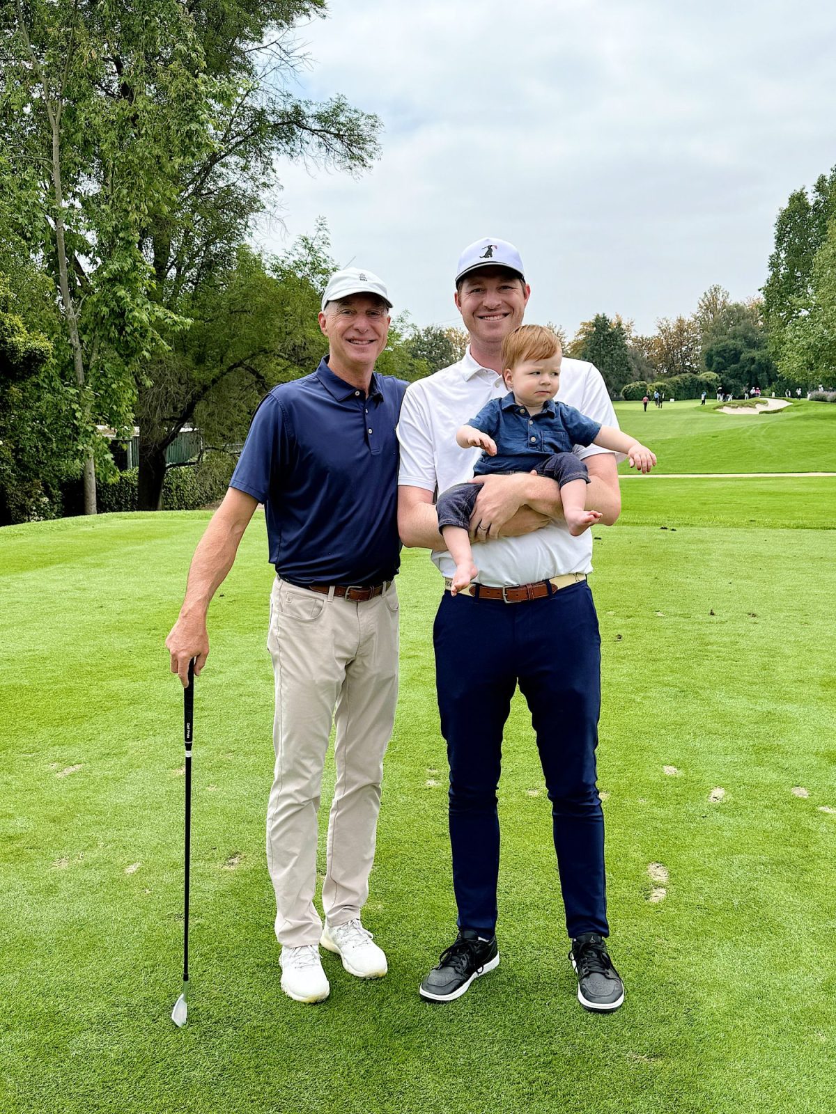 Two men, one holding a baby, stand on a golf course with trees in the background. One man holds a golf club.