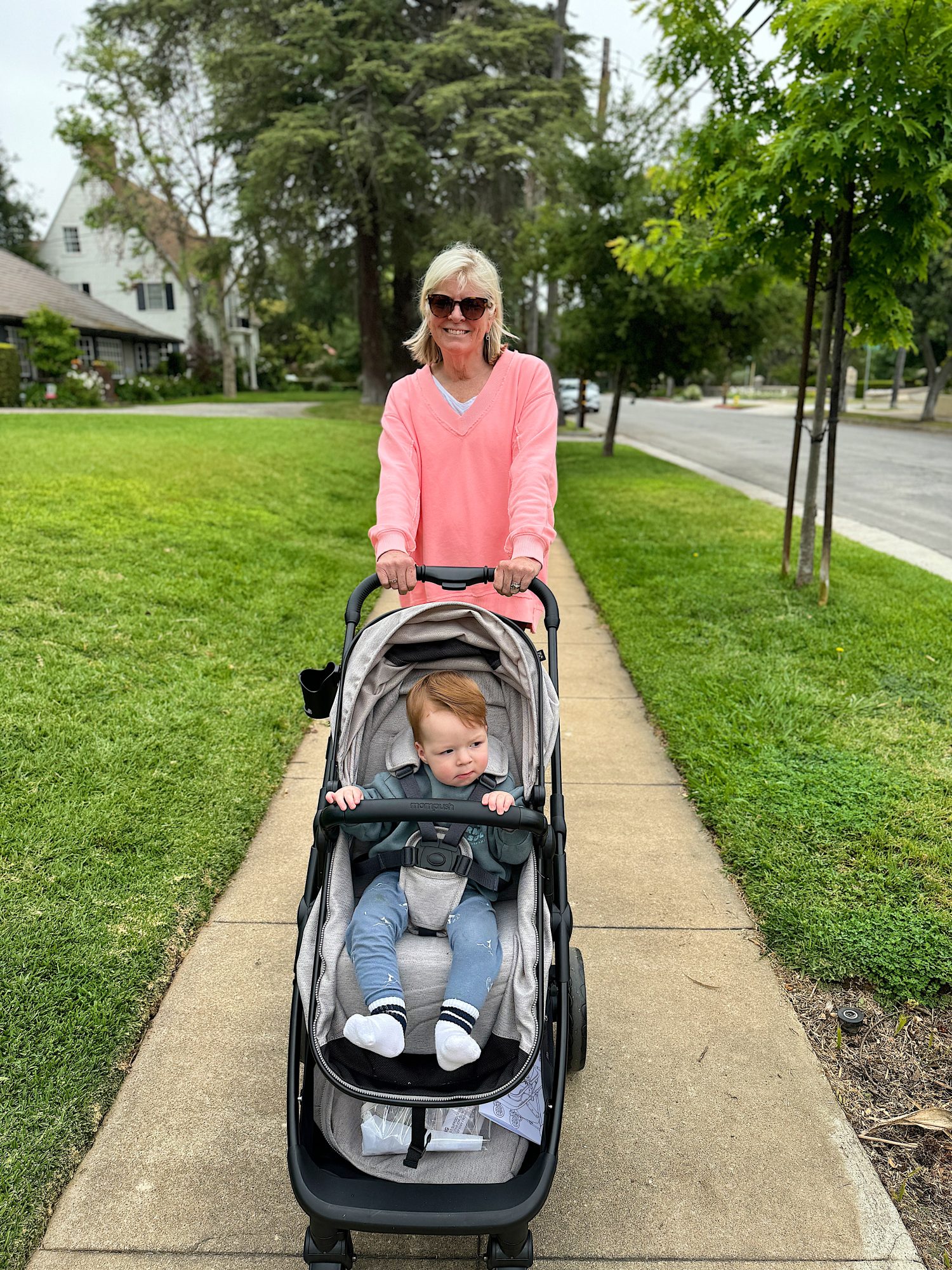 An adult woman wearing sunglasses and a pink sweater pushes a baby in a stroller along a sidewalk. The surrounding area has green grass, trees, and houses.