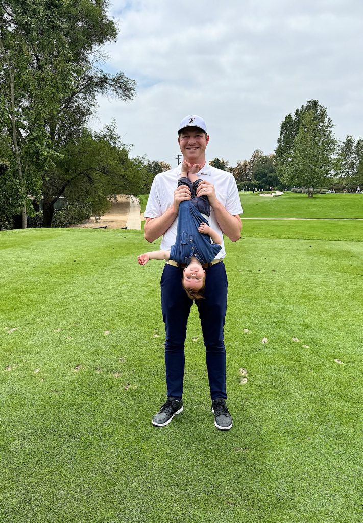 A man in a white shirt and cap is standing on a golf course holding a young child upside down by the legs. The child is laughing and wearing blue overalls. Trees and a golf cart are in the background.