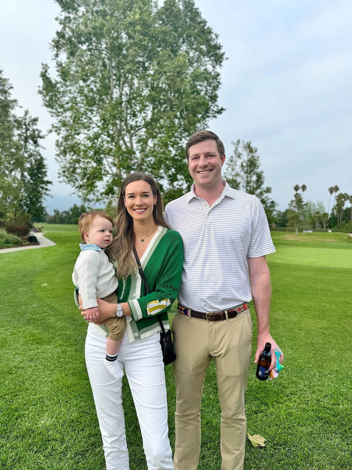 A man and woman stand on a golf course with the woman holding a young child. The man holds a drink and a snack. Trees and grass are visible in the background.