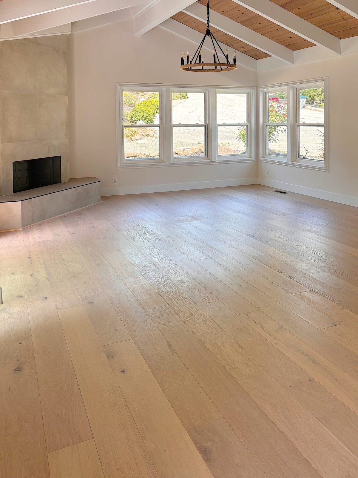 A bright, empty living room with hardwood floors, a large window, a ceiling chandelier, and a modern fireplace on the left side. The ceiling has exposed wooden beams.