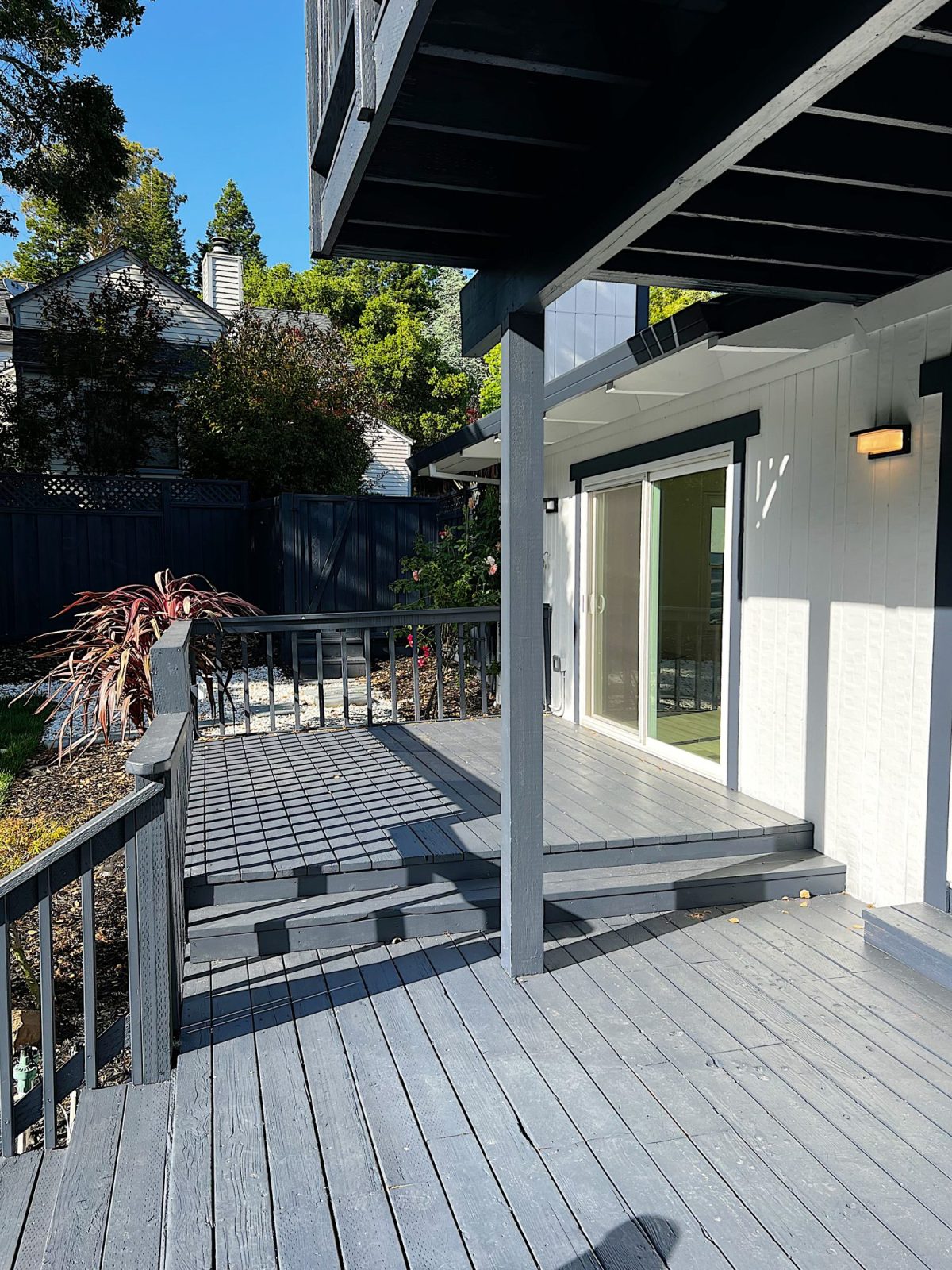 A wooden deck attached to a white house with sliding glass doors, set in a backyard with trees and neighboring houses visible in the background.