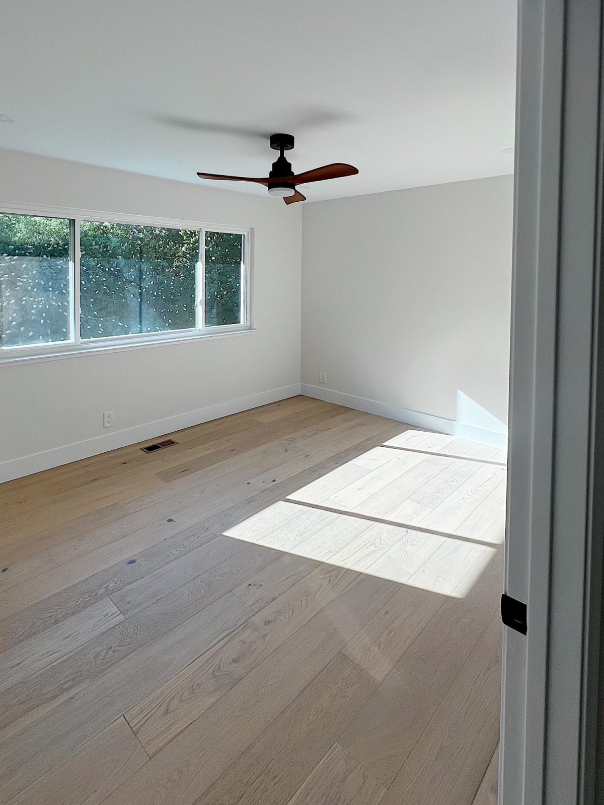 An empty room with a large window, white walls, light wood flooring, and a ceiling fan. Sunlight streams in through the window.