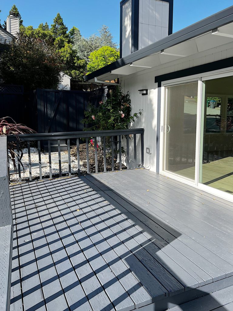A sunlit wooden deck with gray flooring and railings attached to a white house with sliding glass doors leading inside. A small garden area with plants is visible in the background.