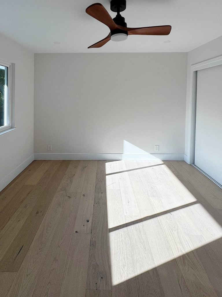Sunlight shines into a minimalist, unfurnished room with wooden floors, white walls, a window on the left, a ceiling fan, and a sliding door on the right.