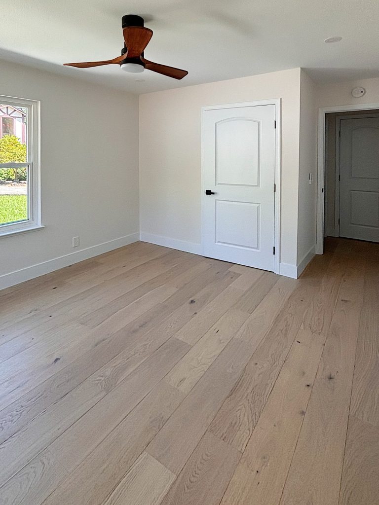 An empty room with light wooden flooring, white walls, a ceiling fan, a window, and two white doors.
