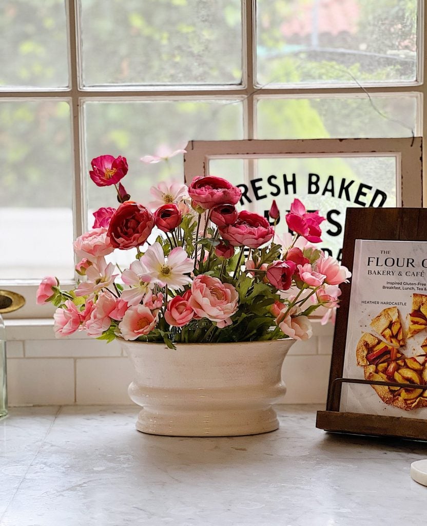 A ceramic pot filled with pink and red flowers sits on a kitchen counter near a window. A bakery sign and a cookbook on a stand are in the background.