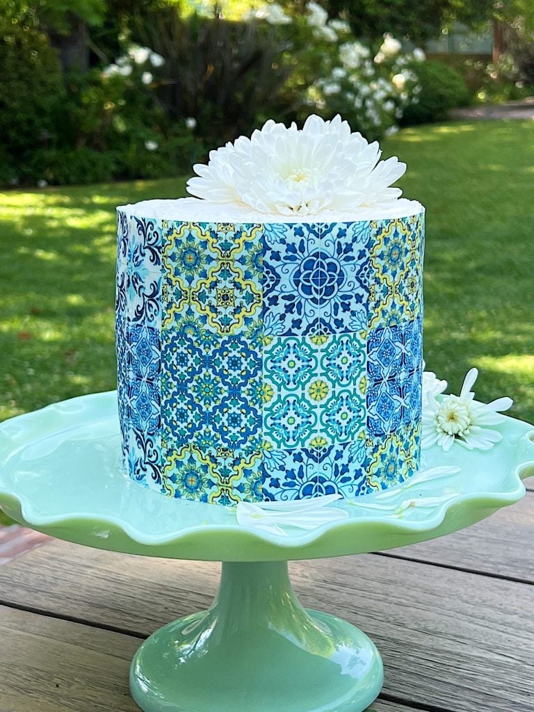 A cake with a blue and white ornate pattern on its exterior, topped with a white chrysanthemum, presented on a light green cake stand in a garden setting.