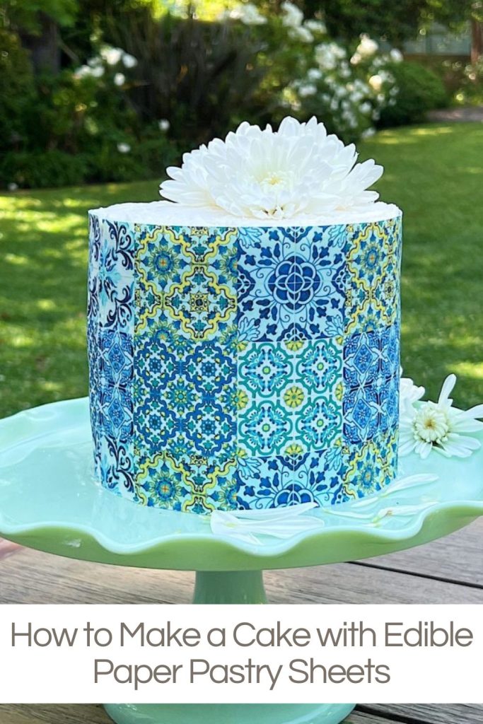 A cake with edible blue and gold patterned pastry sheets, topped with a white flower, displayed on a green plate in a garden setting.