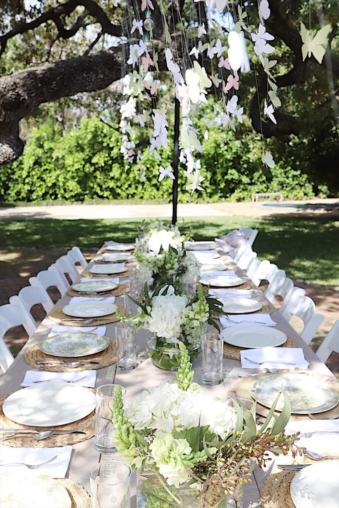 An outdoor dining table is set with white plates, glasses, and floral centerpieces. Paper butterflies hang from above. The scene is surrounded by greenery and trees.