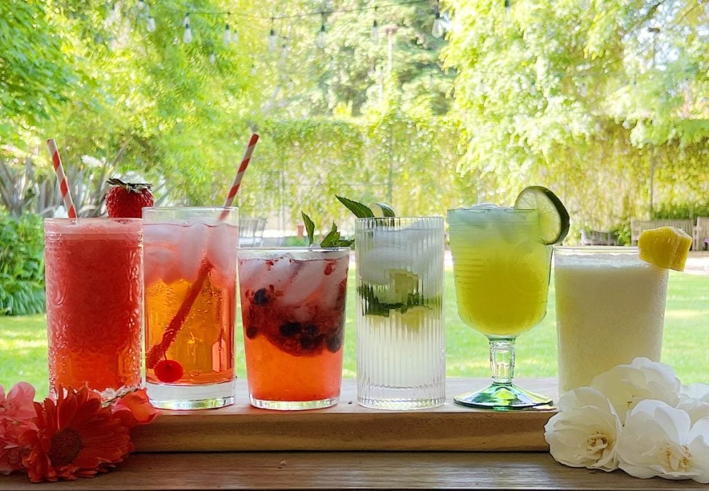 A row of six colorful drinks in various glasses sit on a wooden surface with straws and garnishes, set against a backdrop of green foliage and flowers.