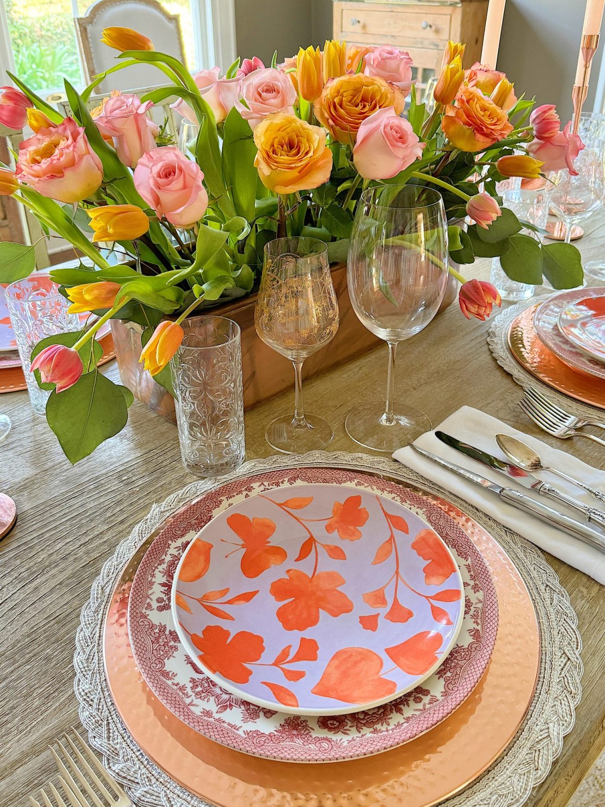 Elegant dining table setting with a floral centerpiece, colorful plates, and crystal glassware, in a room with natural light.