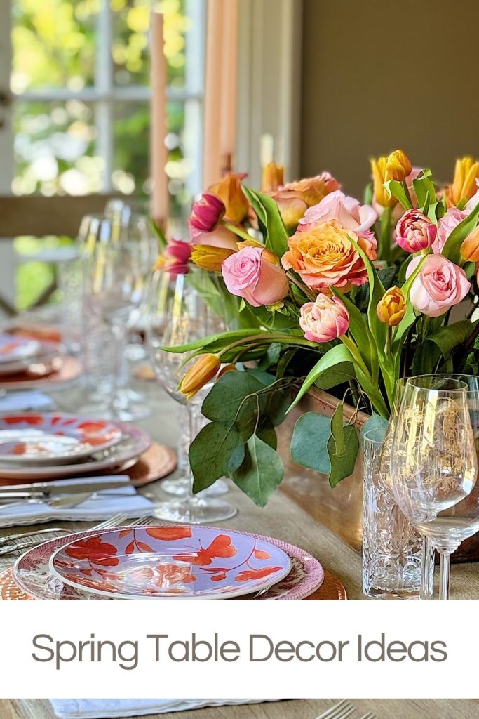 Elegant spring dining table setting with floral plates, crystal glassware, and a bright centerpiece of pink and yellow flowers. Text overlay at the bottom reads: "Spring Table Decor Ideas".