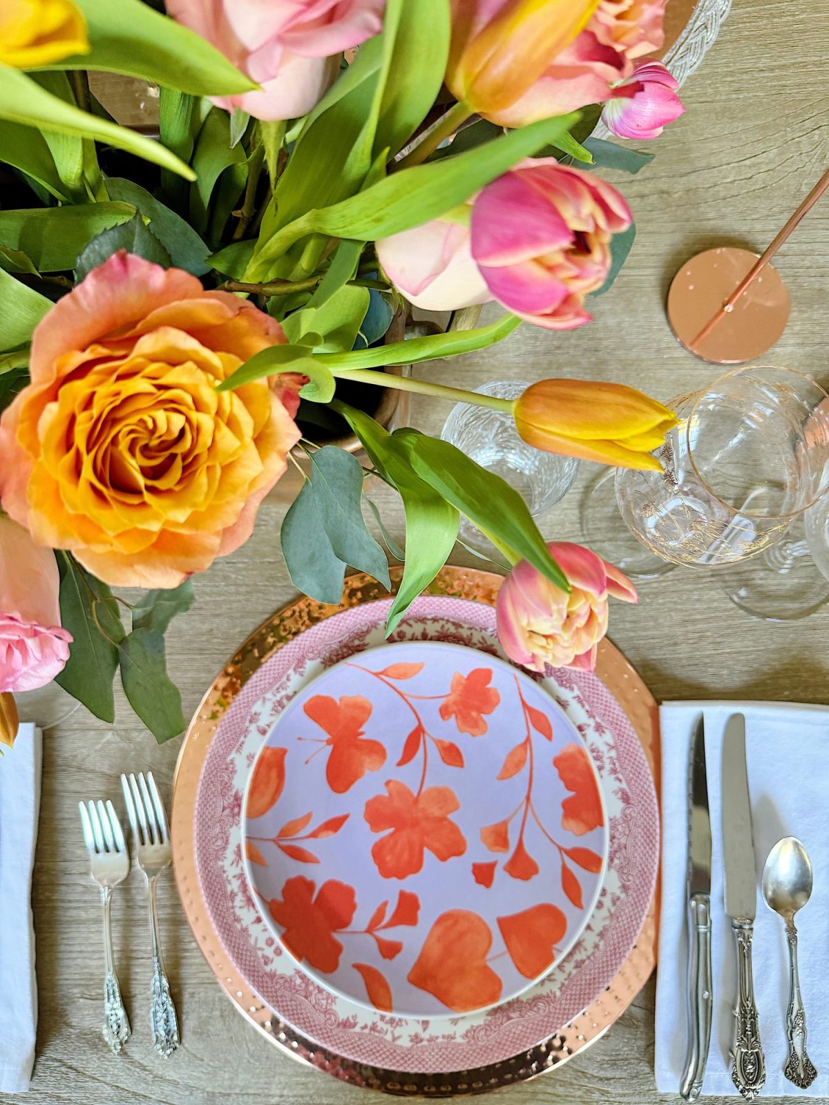 Elegant dining table setting with a floral centerpiece, colorful plates, and crystal glassware, in a room with natural light.