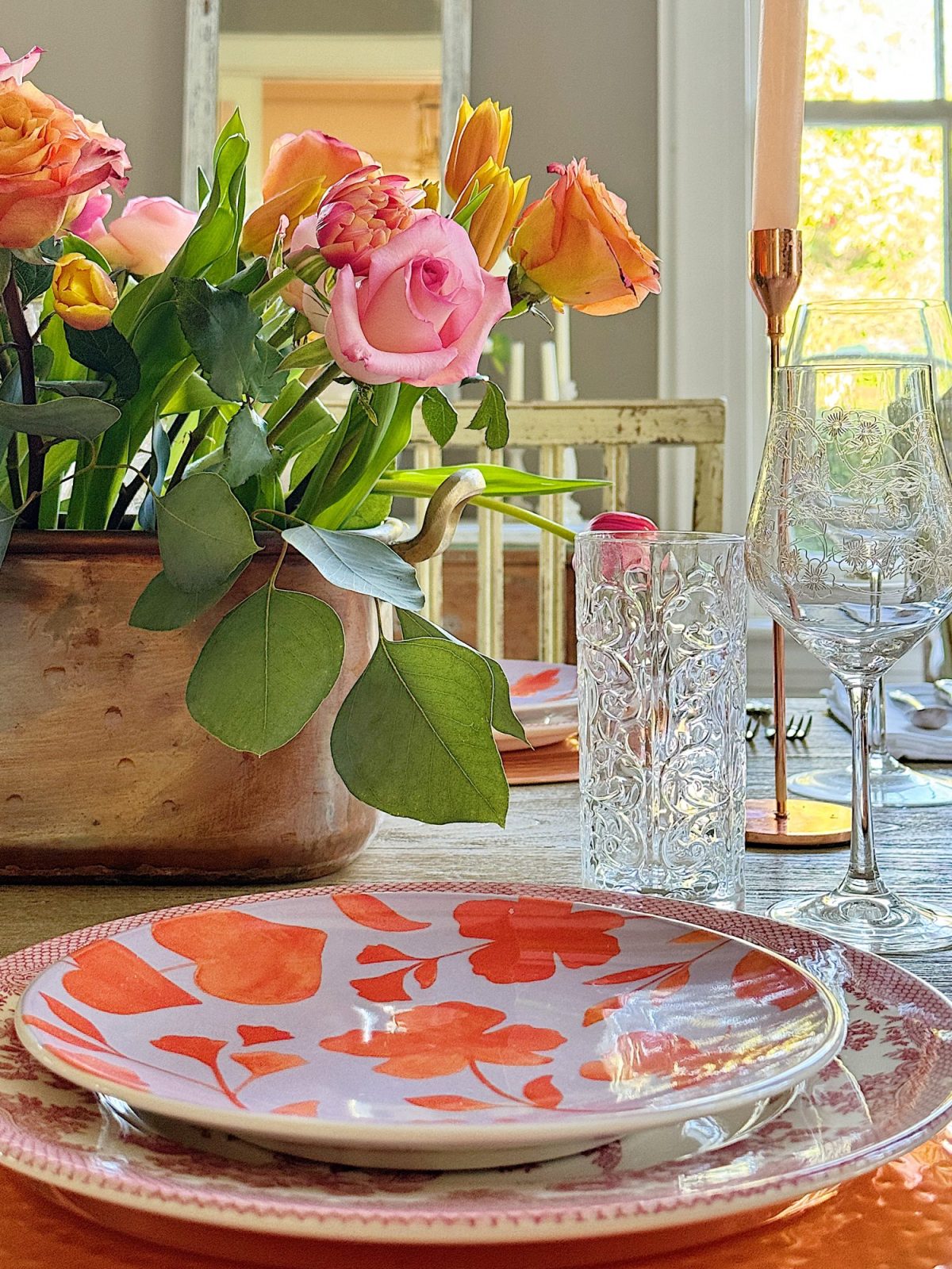 Elegant dining table setting with a floral centerpiece, colorful plates, and crystal glassware, in a room with natural light.