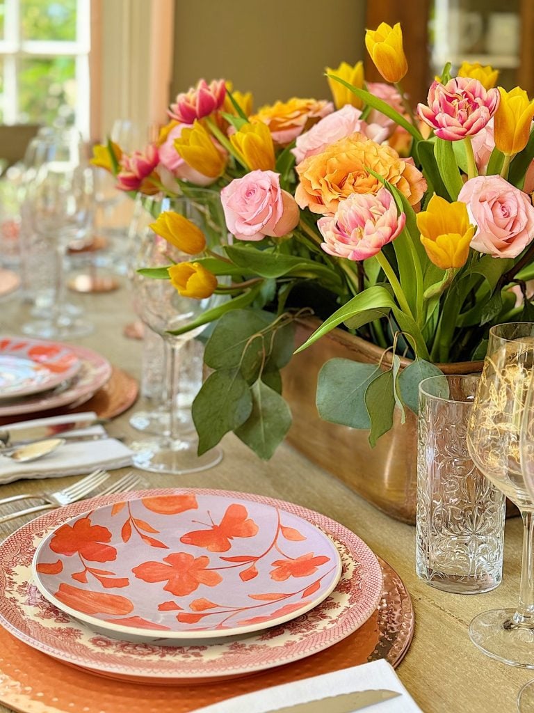 Elegant dining table setting with a floral centerpiece, colorful plates, and crystal glassware, in a room with natural light.
