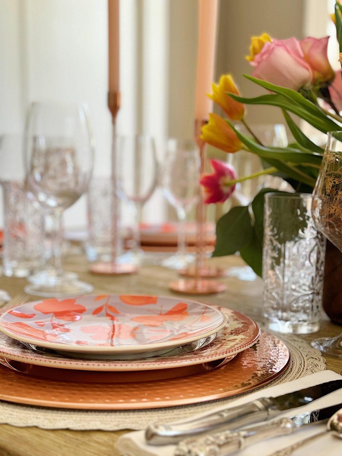 Elegant dining table setting with a floral centerpiece, colorful plates, and crystal glassware, in a room with natural light.
