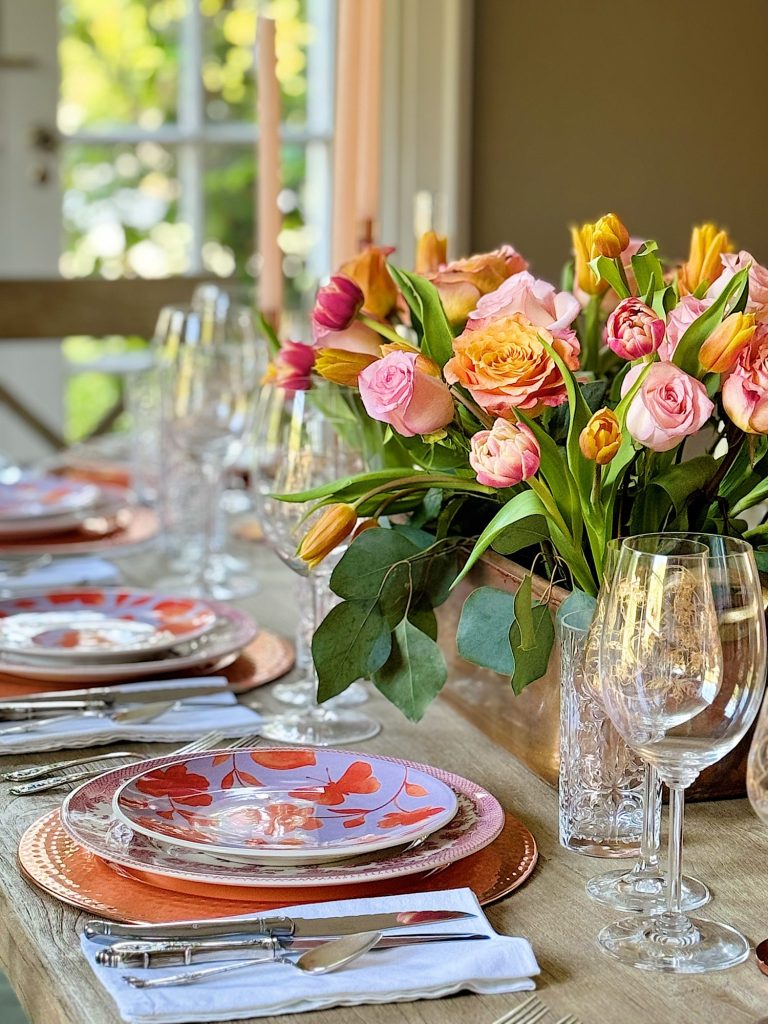 Elegant dining table setting with a floral centerpiece, colorful plates, and crystal glassware, in a room with natural light.