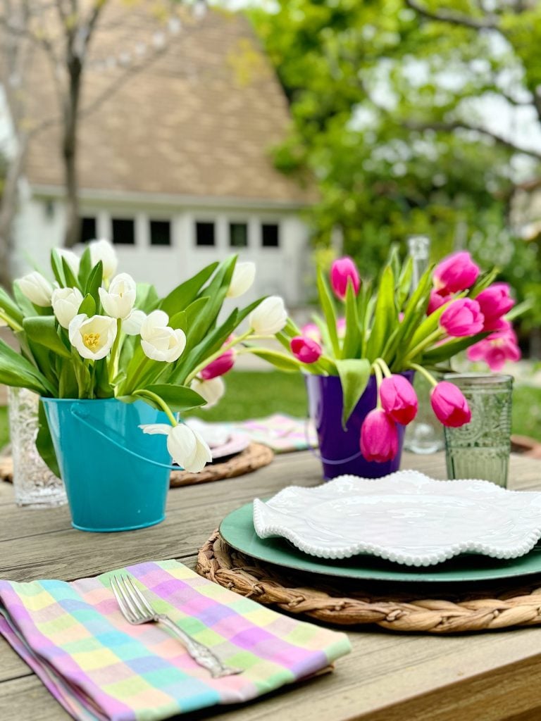 Outdoor table setting featuring colorful tulips in pots, glassware, and white plates on a wooden table with a house in the background.