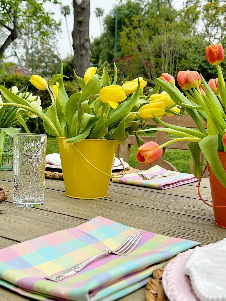 Outdoor dining table set with colorful tulips in pots, checkered napkins, and glassware, focusing on a vibrant, springtime setting.