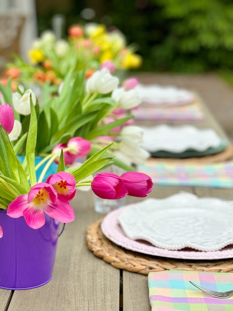 A vibrant bouquet of pink and white tulips in a purple bucket on a table set for a meal, with plates and colorful napkins in the background.