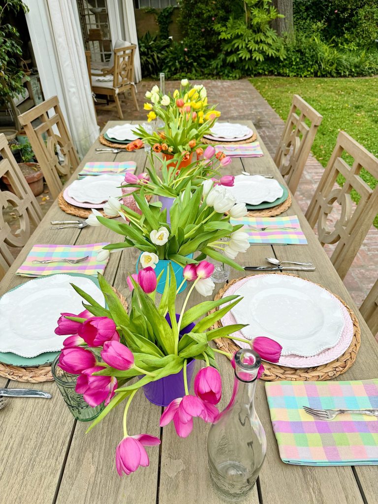 Outdoor dining table set for a meal with floral centerpieces, surrounded by a garden. white plates, colorful napkins, and wooden chairs visible.