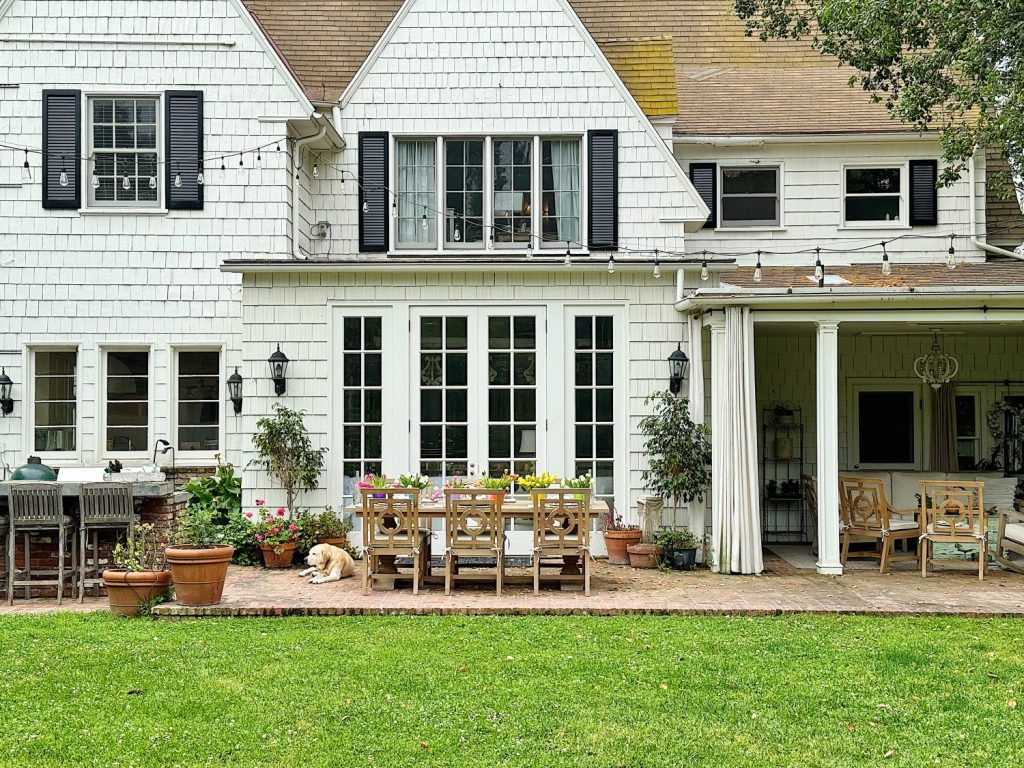 A well-manicured backyard with a large white house featuring french doors. a furnished patio area includes a dining table set and a dog lounging on the grass.