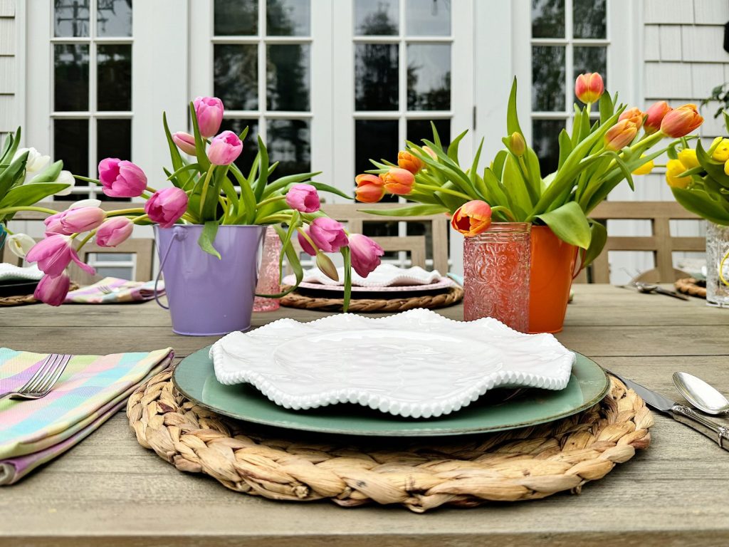 Outdoor dining table set with plates and tulips in colorful pots, with a house in the background.
