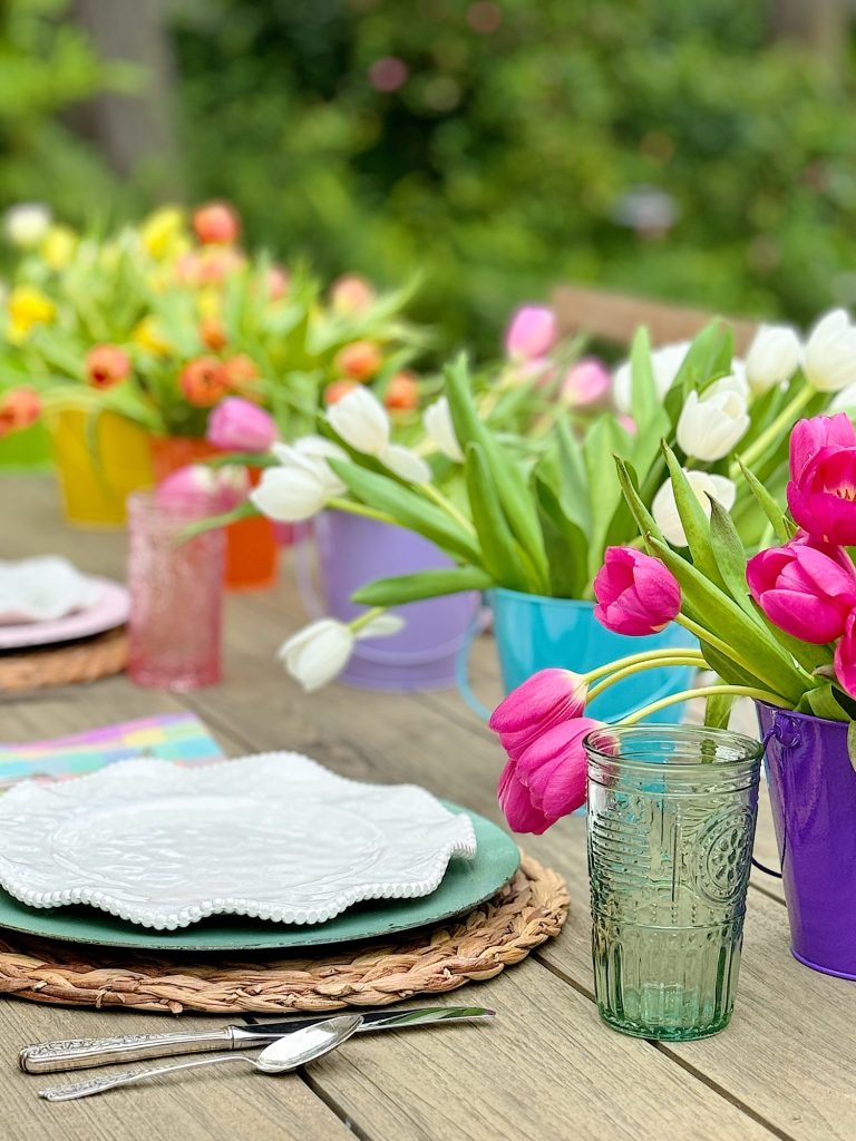 Colorful tulips in buckets on a garden table set for a meal, with plates, utensils, and glasses, focusing on a vibrant, outdoorsy springtime theme.