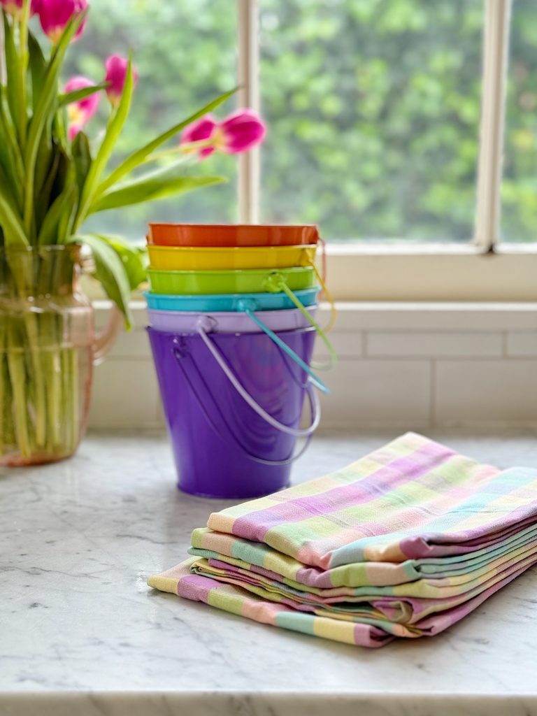 Stacked colorful metal buckets on a marble counter by a window, alongside folded pastel striped cloths and a vase of pink tulips.