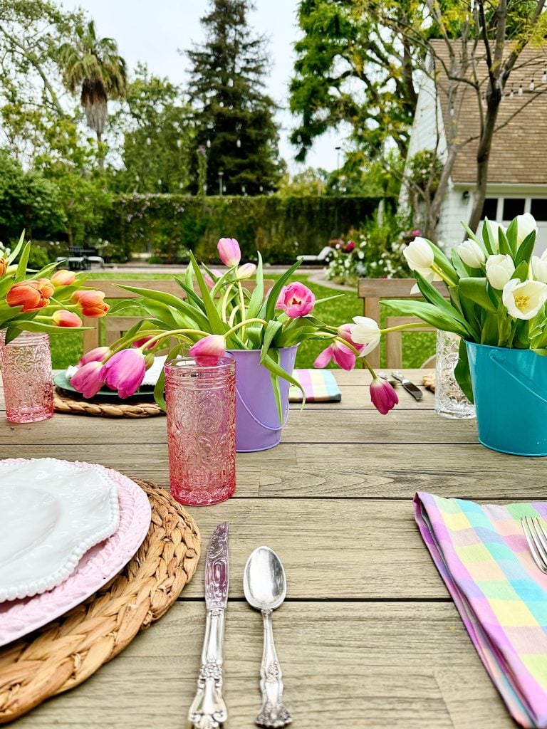 Outdoor dining table set with tulips in colorful pots, plaid tablecloth, and vintage silverware, with a garden view in the background.