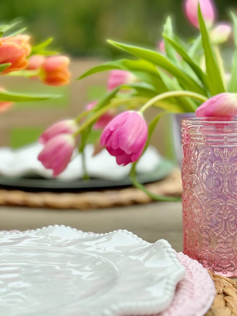 Pink tulips in a glass vase on a table set with white plates and pink placemats, with a blurred garden background.