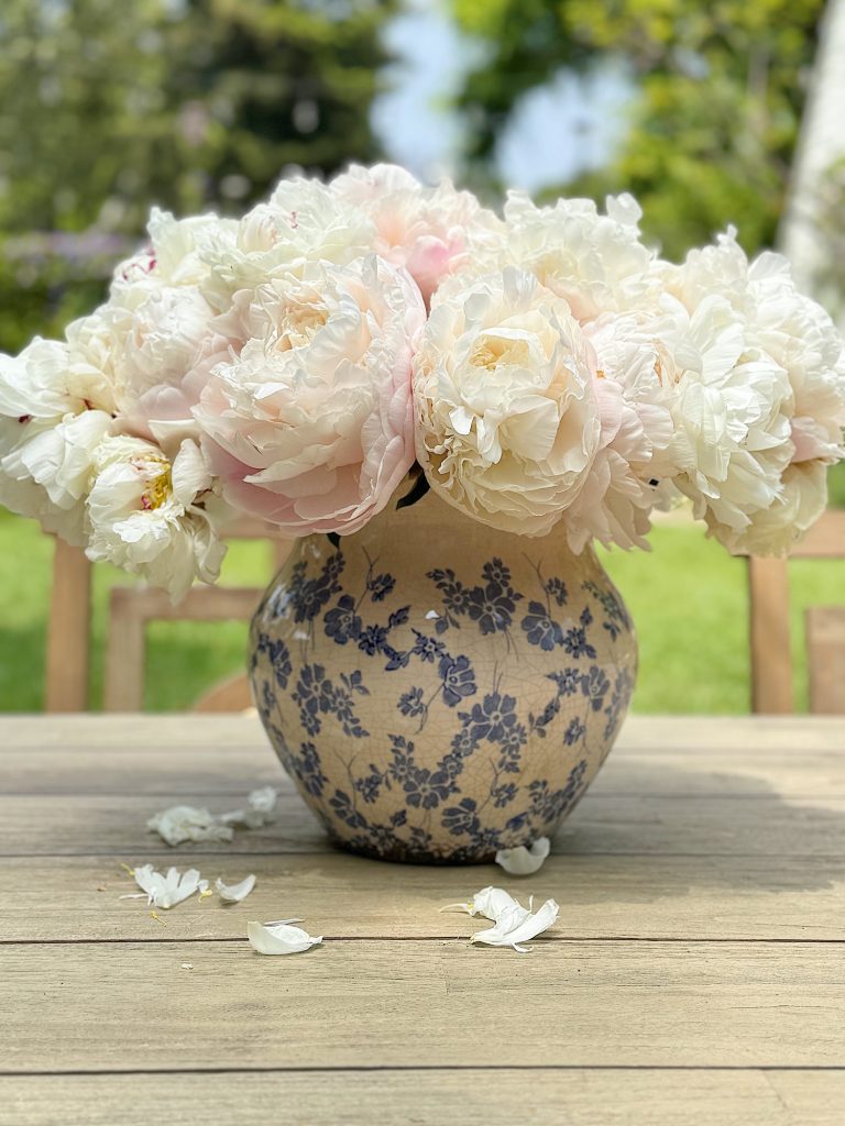 A blue and white floral patterned vase filled with white and light pink peonies sits on a wooden table outside, with a few fallen petals scattered around.