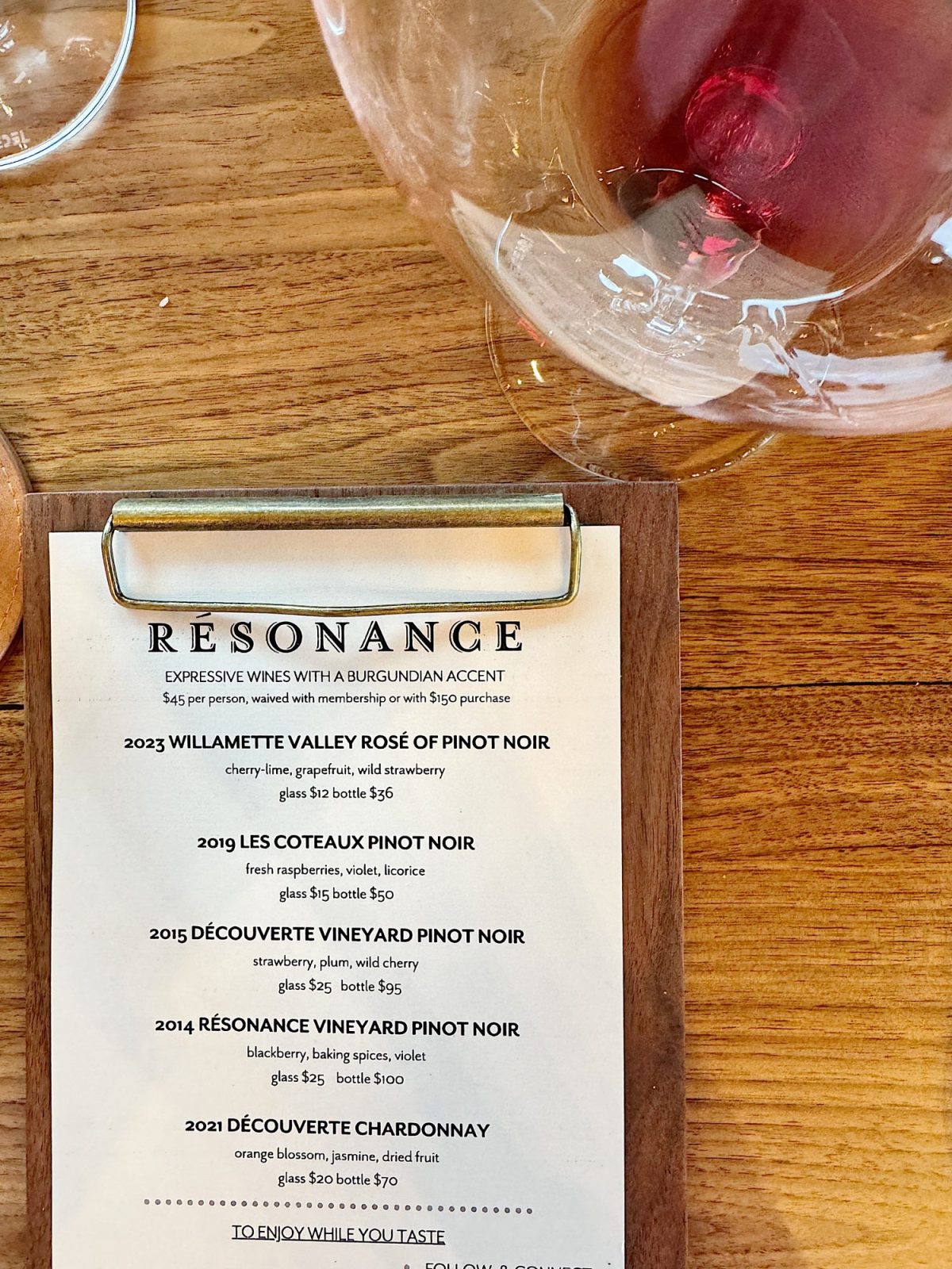 Wine list on a wooden table featuring selections from different vintages, beside an empty wine glass with a rose petal.
