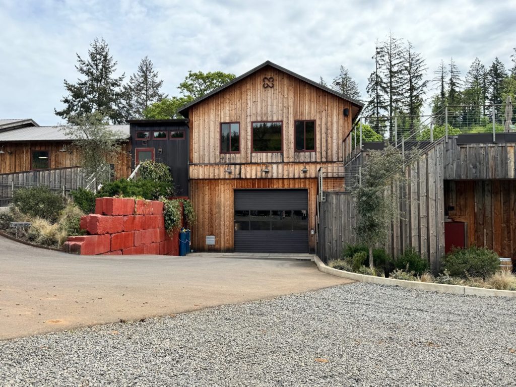 A rustic barn-style winery with a wooden facade, garage door, and a red block structure in the driveway, surrounded by trees and gravel.