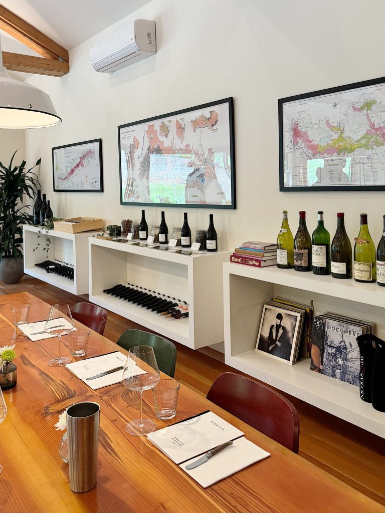 Modern dining room with a wooden table set for four, white shelves with books and wine bottles, wall-mounted maps, and an indoor plant.