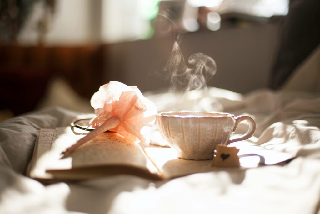 A cozy morning scene with a steaming cup of tea, an open book, and glasses on a sunlit bed, evoking a peaceful ambiance.